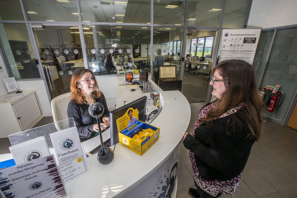 Image shows a woman behind the reception desk at Glamorgan Archives, speaking to another woman. Credit Archives and Records Council Wales