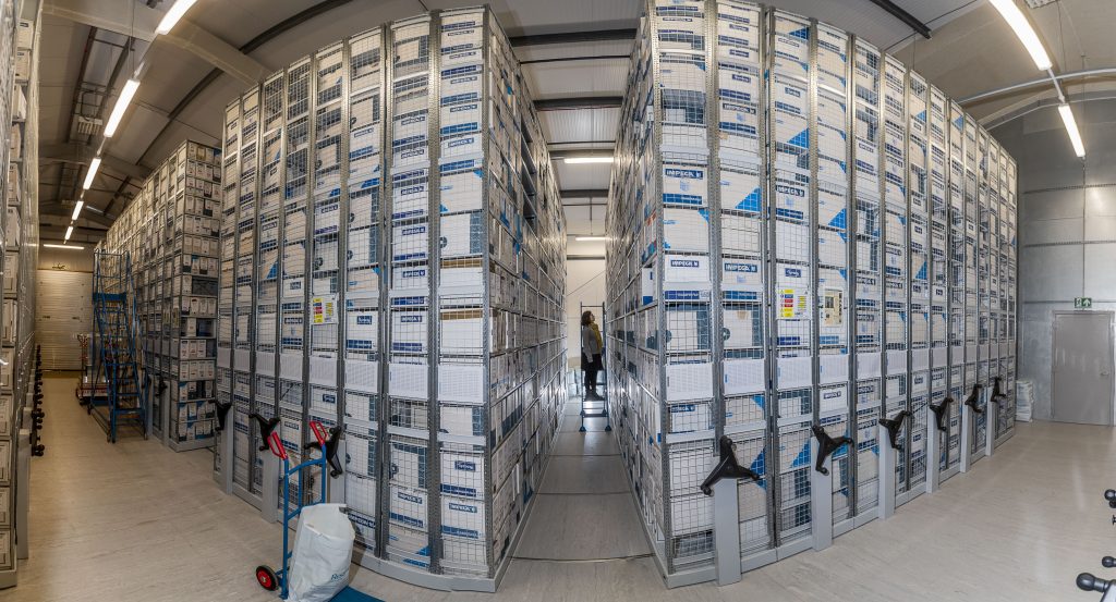 Image shows large archive stacks with a person walking past in the distance. Credit Archives and Records Council Wales
