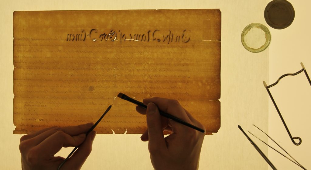 A document and conservation tools is lit from behind, we can see a pair of hands working on the document. Image courtesy of Mark O'Neill